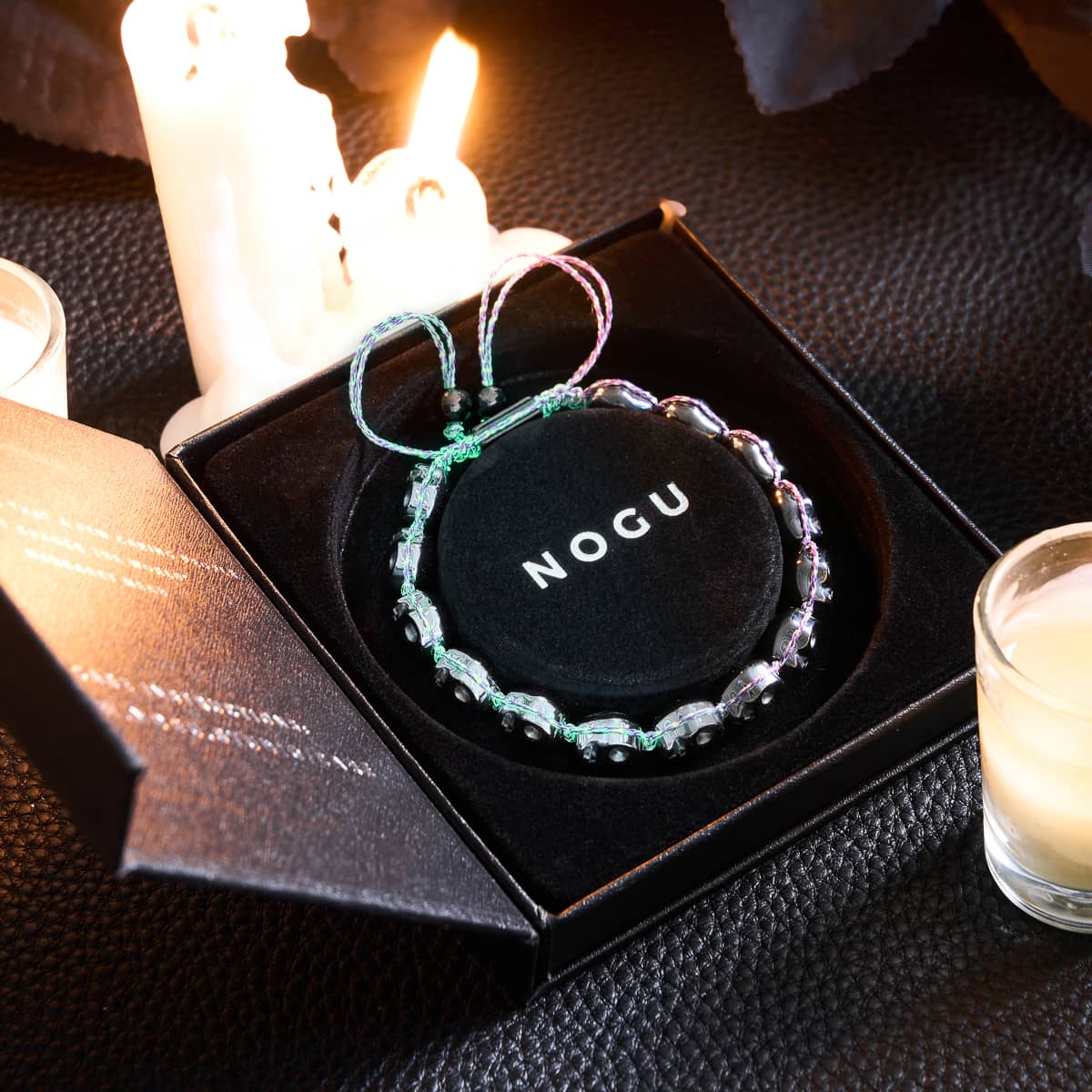 Witching Hour gunmetal skull bead macrame bracelet with violet GlowWeave cord displayed in black NOGU box with candles